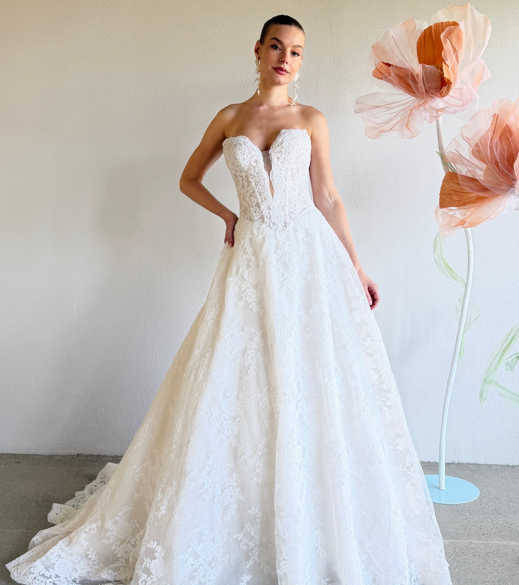 Woman in a white strapless lace wedding dress standing next to large floral arrangements.