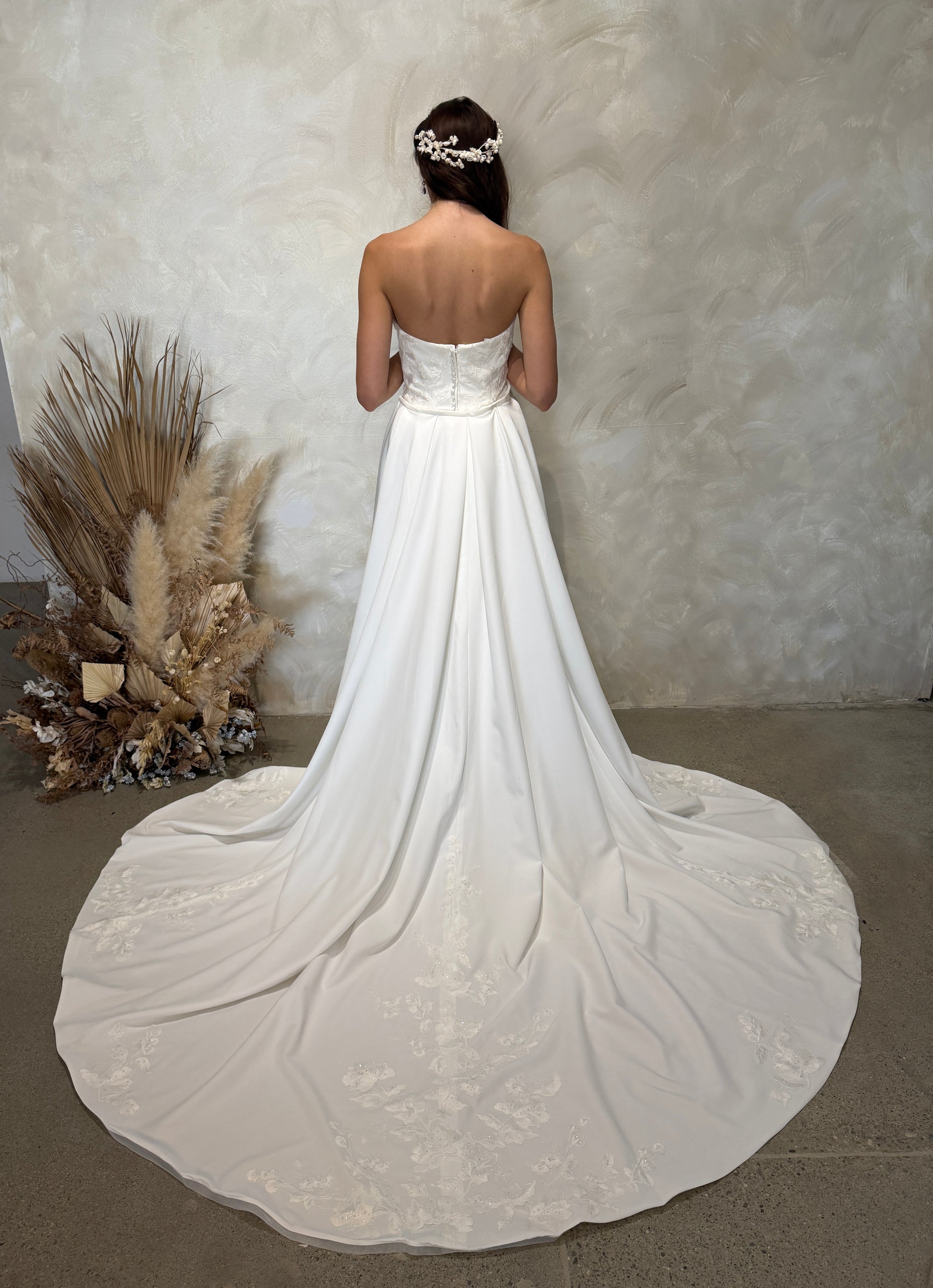 Back view of the Aveline fitted strapless wedding dress with floral lace appliqué and detachable draped train panels