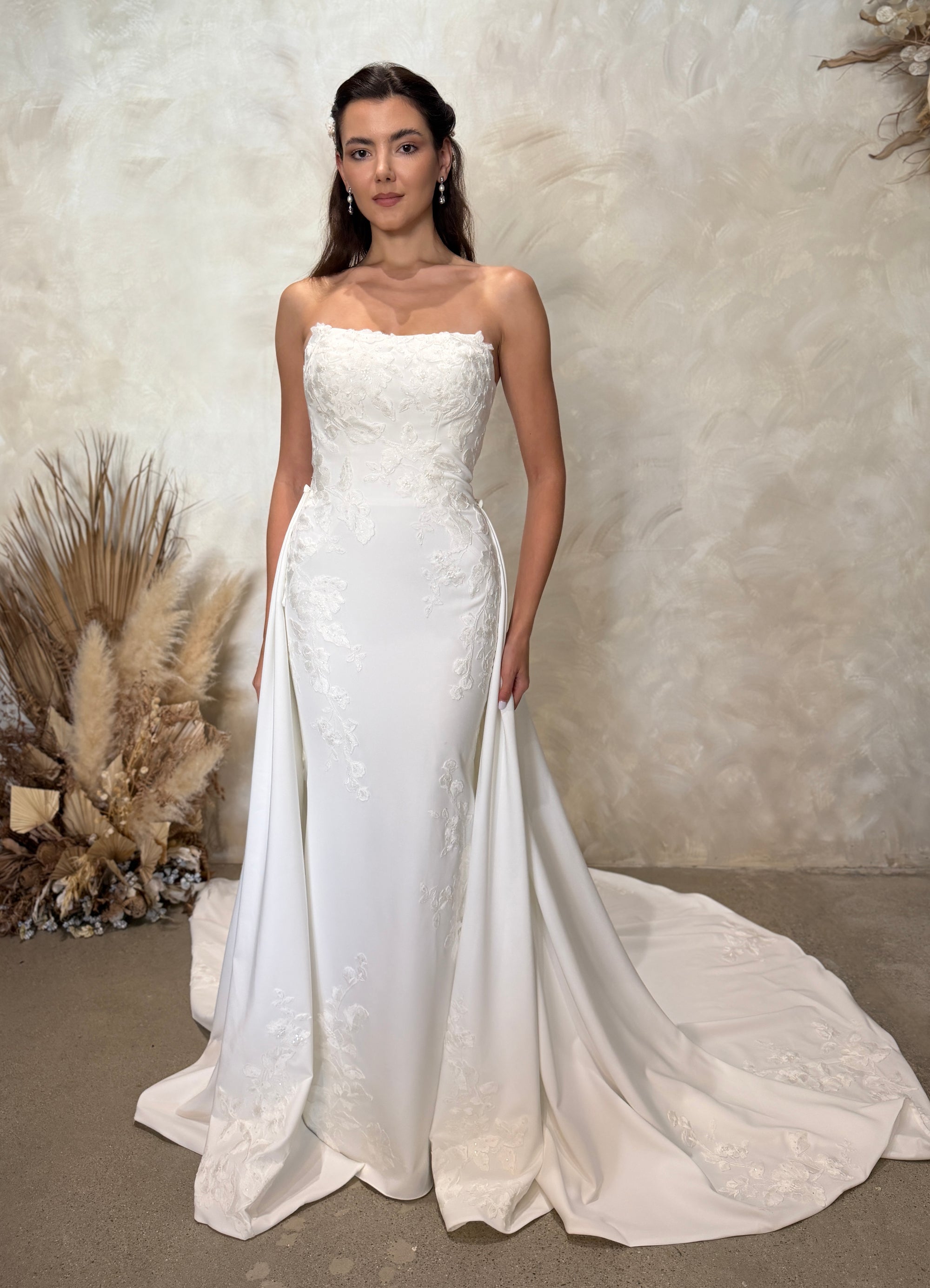 Front view of the Aveline fitted strapless wedding dress with floral lace appliqué and detachable draped train panels